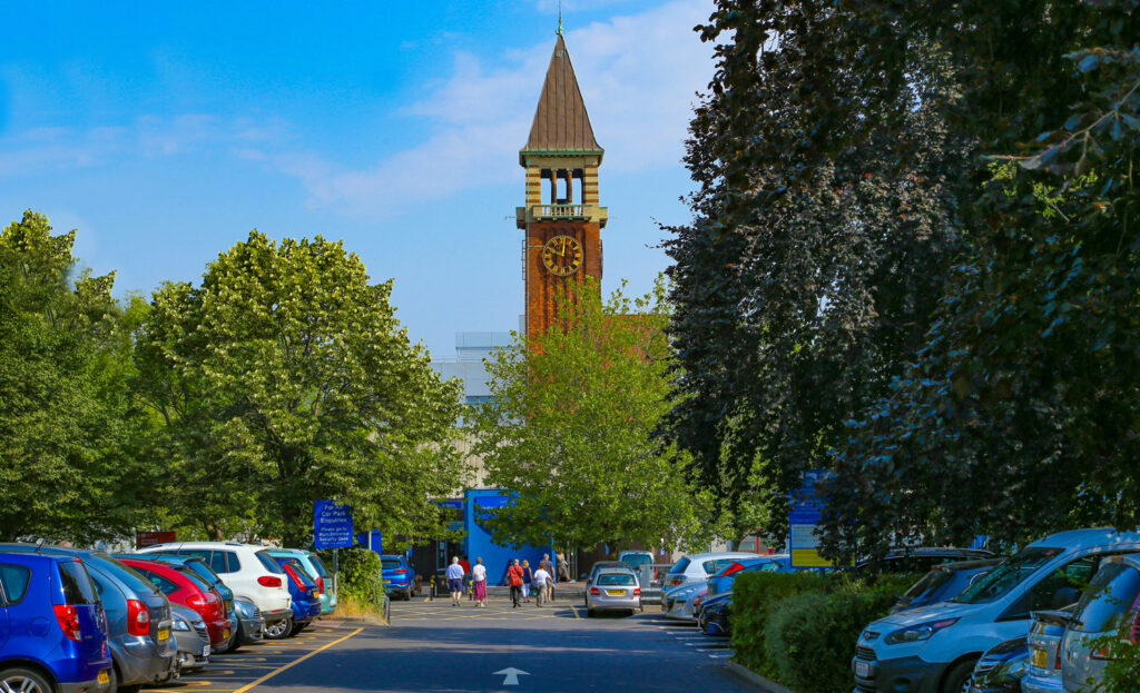 A hospital car park with people walking towards the main entrance. The Clock Tower of the building can be seen behind the trees.