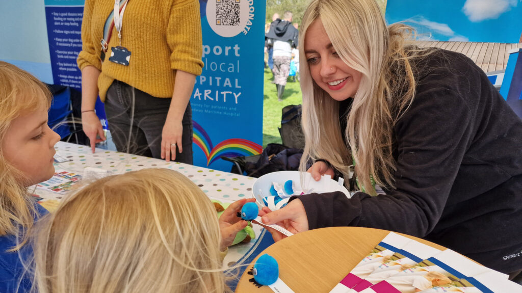 A volunteer hands a blue fuzzy bug to a child at an outdoor fair